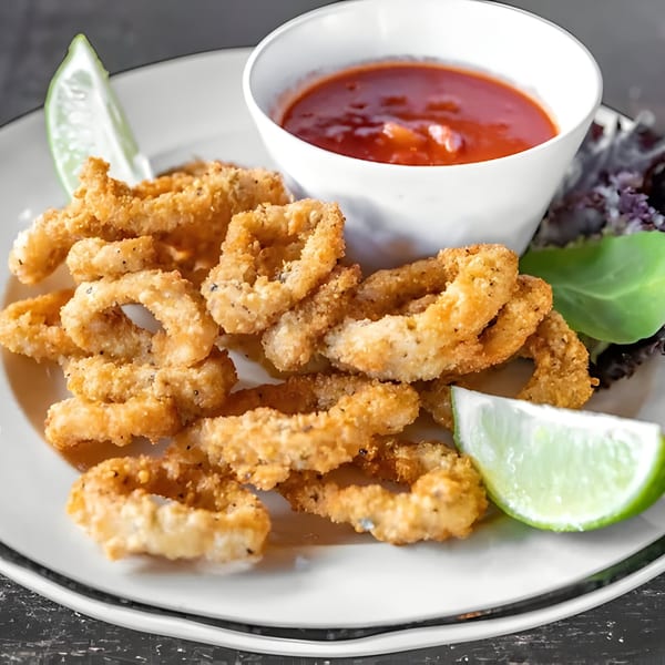 Crispy fried calamari rings with a side of red dipping sauce and lime wedges on a white plate.