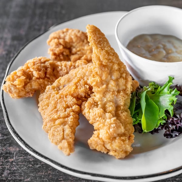 Crispy chicken tenders with a side of dipping sauce and fresh greens on a white plate.