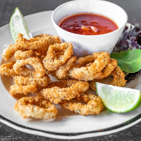 Crispy fried calamari rings with a side of red dipping sauce, lime wedges, and lettuce.