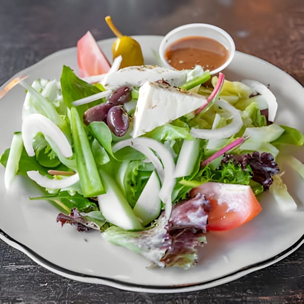 A fresh side salad with mixed greens, tomatoes, onions, olives, feta cheese, and a side of dressing.